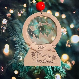 May include: A wooden Christmas ornament shaped like a snow globe with a white silhouette of Santa Claus and reindeer flying through the air. The ornament has a banner that reads "2021 Merry Christmas".