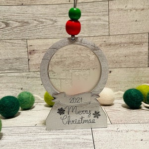 May include: A white wooden Christmas ornament with a frosted glass globe, a red and green beaded string, and the text "2021 Merry Christmas".