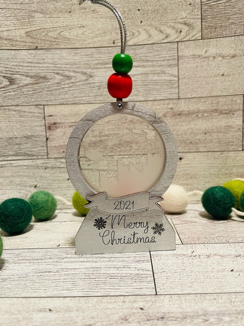 May include: A white wooden Christmas ornament with a frosted glass globe, a red and green beaded string, and the text "2021 Merry Christmas".