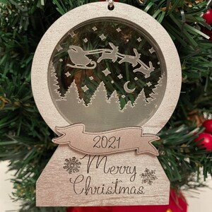 May include: A silver Christmas ornament shaped like a snow globe with a scene of Santa Claus flying in his sleigh with reindeer. The ornament has a banner that says "2021 Merry Christmas".