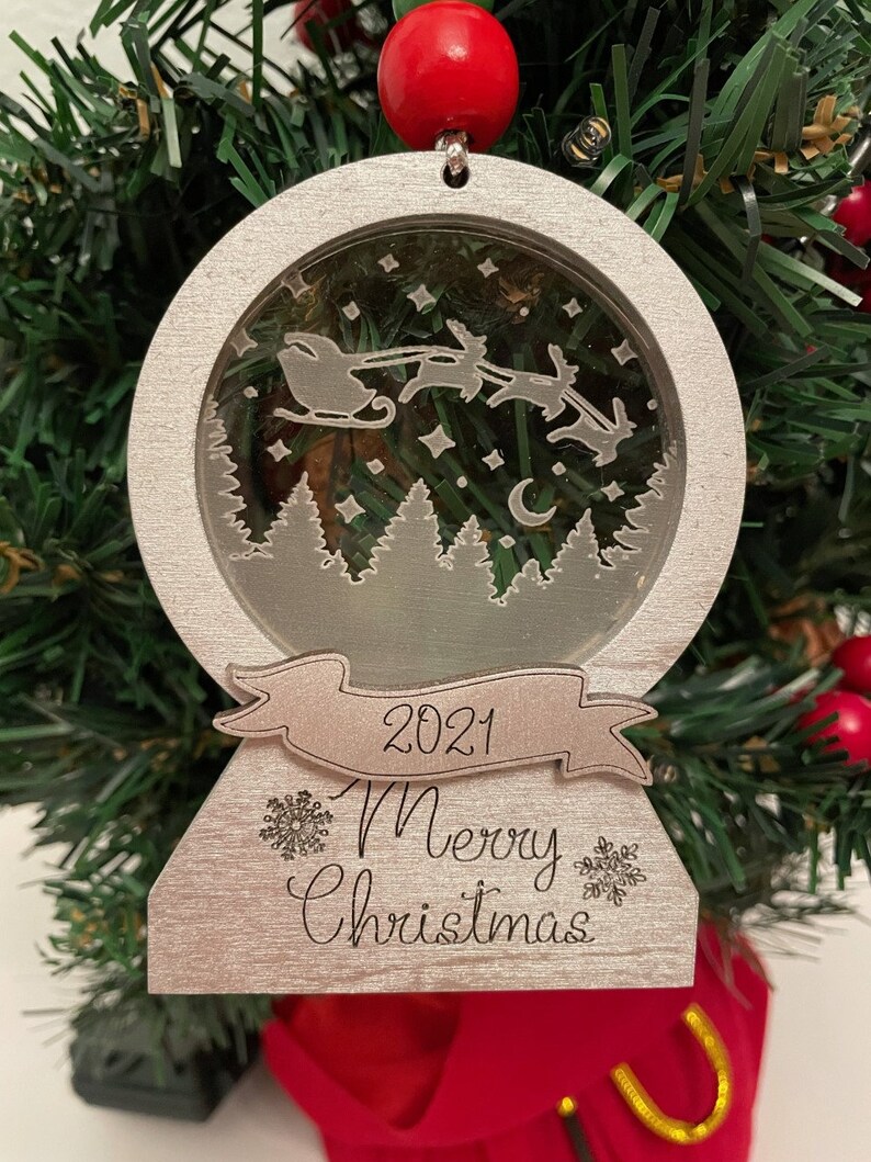 May include: A silver Christmas ornament shaped like a snow globe with a scene of Santa Claus flying in his sleigh with reindeer. The ornament has a banner that says "2021 Merry Christmas".
