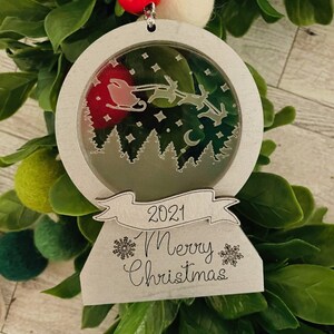 May include: A silver Christmas ornament with a snow globe design. The globe features a silhouette of Santa Claus flying with reindeer over a forest. The ornament has a banner with the text "2021 Merry Christmas" and snowflake designs.