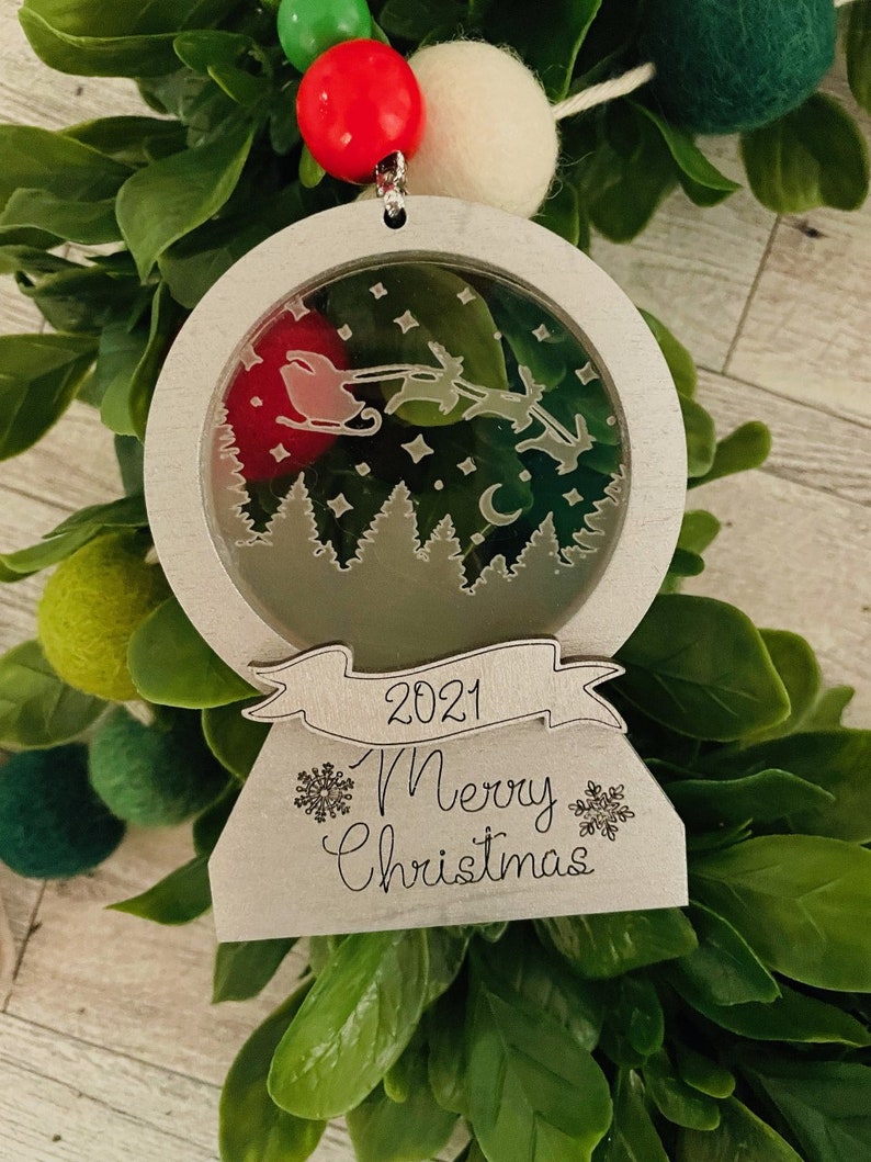 May include: A silver Christmas ornament with a snow globe design. The globe features a silhouette of Santa Claus flying with reindeer over a forest. The ornament has a banner with the text "2021 Merry Christmas" and snowflake designs.
