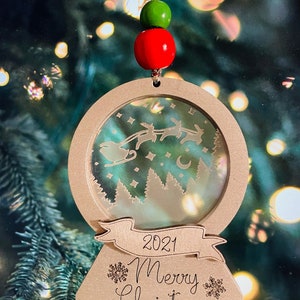 May include: A wooden Christmas ornament shaped like a snow globe with a scene of Santa Claus flying in his sleigh with reindeer. The ornament has a banner with the text "2021 Merry Christmas" and is decorated with snowflakes.