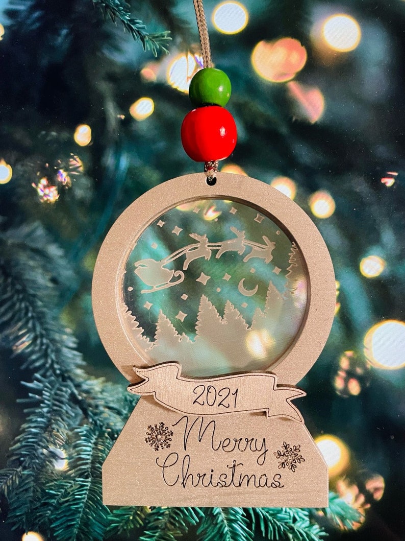 May include: A wooden Christmas ornament shaped like a snow globe with a scene of Santa Claus flying in his sleigh with reindeer. The ornament has a banner with the text "2021 Merry Christmas" and is decorated with snowflakes.