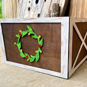 May include: A wooden box with a white distressed frame and a dark brown center panel. A green wreath with red berries is affixed to the front. The right side features an X-shaped design.