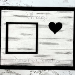 Puede incluir: Un letrero de madera blanco con borde negro y el texto "Oh baby! We can't wait to meet you!" El letrero tiene un recorte de corazón negro y un marco negro para una foto.