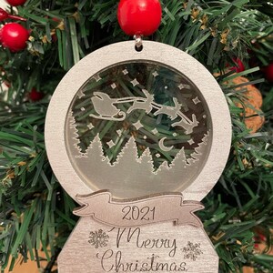 May include: A silver Christmas ornament shaped like a snow globe with a scene of Santa Claus flying in his sleigh pulled by reindeer. The ornament has a banner that reads "2021 Merry Christmas".
