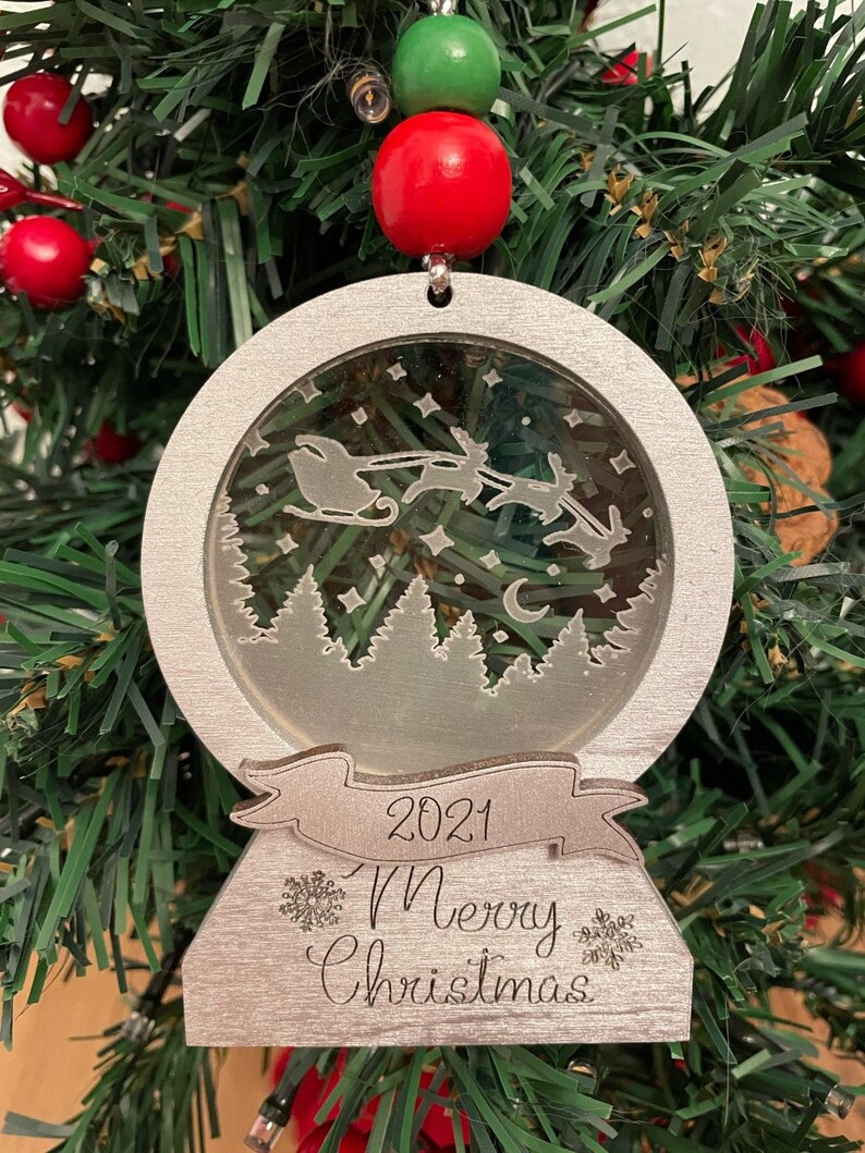 May include: A silver Christmas ornament shaped like a snow globe with a scene of Santa Claus flying in his sleigh pulled by reindeer. The ornament has a banner that reads "2021 Merry Christmas".