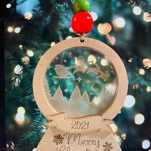 May include: A wooden Christmas ornament shaped like a snow globe with a scene of Santa Claus flying through the air with reindeer. The ornament has a banner that says "2021 Merry Christmas".