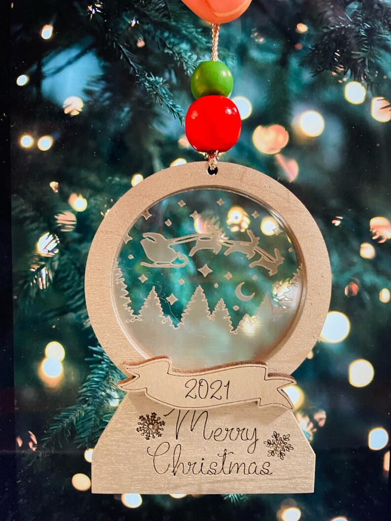 May include: A wooden Christmas ornament shaped like a snow globe with a scene of Santa Claus flying through the air with reindeer. The ornament has a banner that says "2021 Merry Christmas".
