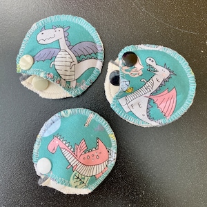 May include: Three round, teal fabric items with a dragon print. Each has a different dragon design in white and pink, with snap closures. The edges are stitched with white thread, and the background has a light green and white pattern.