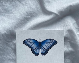 May include: A blue butterfly with dark blue markings on its wings is drawn on a white card.