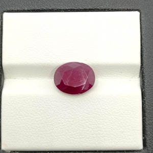 Può includere: Una gemma di rubino sfaccettata di forma ovale. Il rubino è di un colore rosso intenso e ricco, posto su uno sfondo bianco all'interno di un espositore nero. Le sfaccettature della gemma catturano la luce, creando un effetto scintillante.