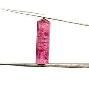 May include: A vibrant pink tourmaline crystal with a columnar, elongated shape and a naturally faceted termination point. The gemstone displays a translucent quality with visible internal inclusions and vertical striations along its length.