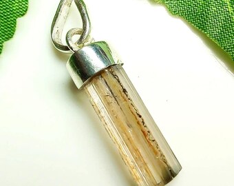 Puede incluir: Un colgante con una gema cilíndrica transparente con inclusiones marrones y negras, engastada en una tapa y un asa de metal plateado. La gema tiene un aspecto natural y texturizado. El colgante está diseñado para ser usado como collar.