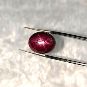 Puede incluir: Una gema pulida, de forma ovalada, de color rojo intenso, con un efecto de estrella, sujeta con pinzas plateadas. La gema tiene una superficie lisa y reflectante, que muestra un patrón de estrella de seis rayos. El fondo es blanco suave y texturizado.