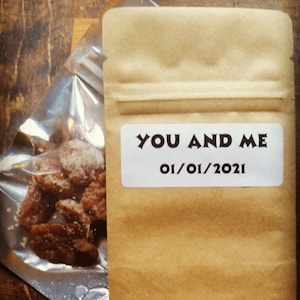 以下が含まれることがあります： 白いラベルに「YOU AND ME 01/01/2021」と書かれた茶色の紙袋。袋は木製の表面に置かれ、砂糖漬けのペカンナッツが入った透明なビニール袋の隣にある。