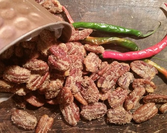 Puede incluir: Primer plano de un bol de mitades de nueces de pecán cubiertas con un glaseado de azúcar moreno. Las nueces de pecán están esparcidas sobre una superficie de madera con chiles rojos y verdes.