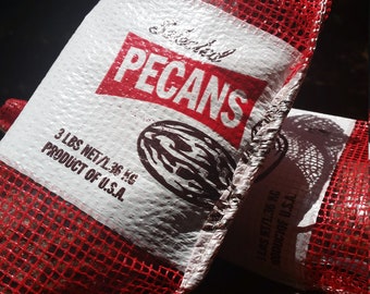 Puede incluir: Una bolsa de malla roja llena de nueces de pecán. La bolsa tiene una etiqueta blanca que dice "Selected Pecans" y "3 lbs net/1.36 kg Producto de EE. UU."