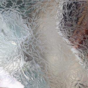 May include: Close-up of a clear glass surface with a detailed, frost-like pattern. The intricate design resembles delicate, feathery formations, creating a textured, natural aesthetic. The glass reflects light, enhancing the visual depth of the design.