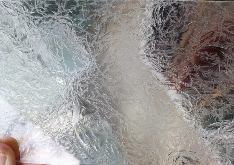 May include: Close-up of a clear glass surface with a detailed, frost-like pattern. The intricate design resembles delicate, feathery formations, creating a textured, natural aesthetic. The glass reflects light, enhancing the visual depth of the design.