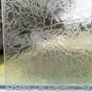 May include: A close-up of a clear glass pane with a detailed frost pattern. The frost creates a delicate, intricate design resembling branches and leaves, with a blurred background visible through the glass.