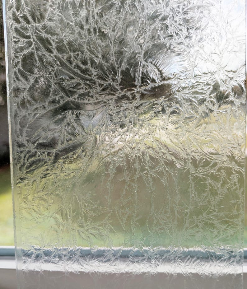 May include: A close-up of a clear glass pane with a detailed frost pattern. The frost creates a delicate, intricate design resembling branches and leaves, with a blurred background visible through the glass.