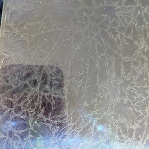 May include: A clear, rectangular glass panel with a frosted, crystallized pattern. The design resembles delicate, branching ice formations. The panel's surface reflects light, creating a subtle, iridescent effect. The overall aesthetic is reminiscent of winter or cold weather.