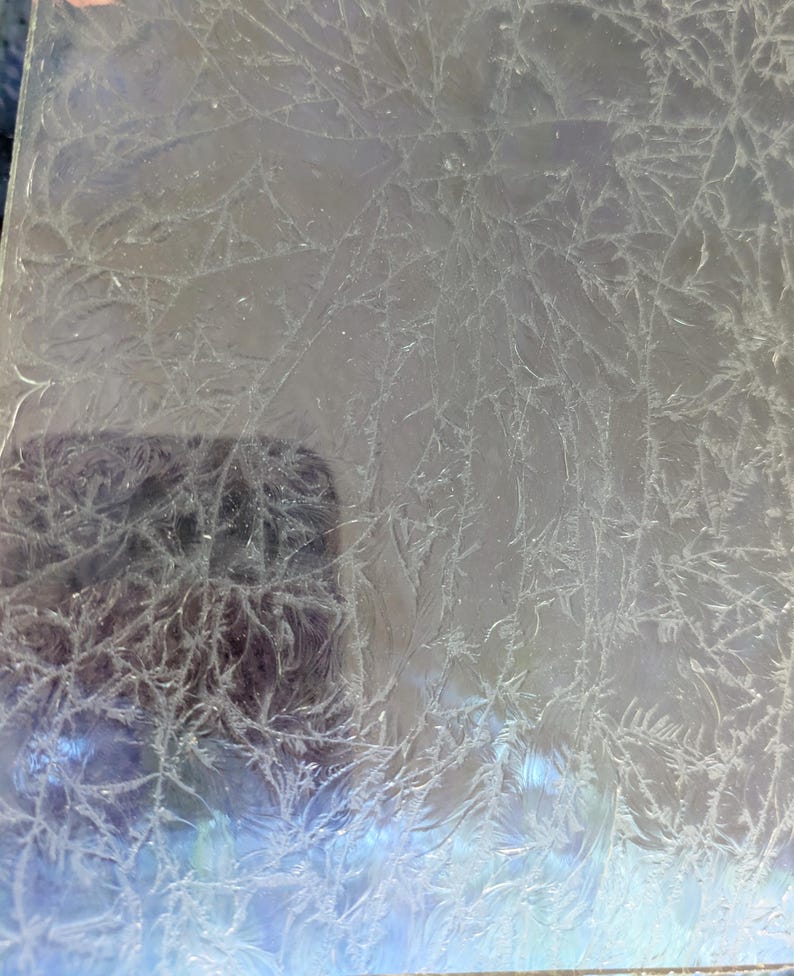 May include: A clear, rectangular glass panel with a frosted, crystallized pattern. The design resembles delicate, branching ice formations. The panel's surface reflects light, creating a subtle, iridescent effect. The overall aesthetic is reminiscent of winter or cold weather.