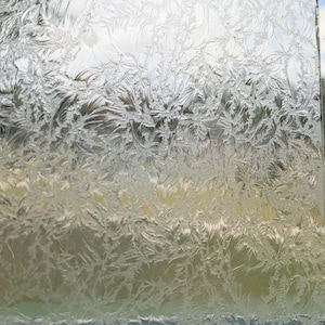May include: A clear glass panel with a frosted, textured surface. The intricate pattern resembles delicate ice crystals or frost, with a mix of light and shadow. The background shows a glimpse of the sky and other colors.