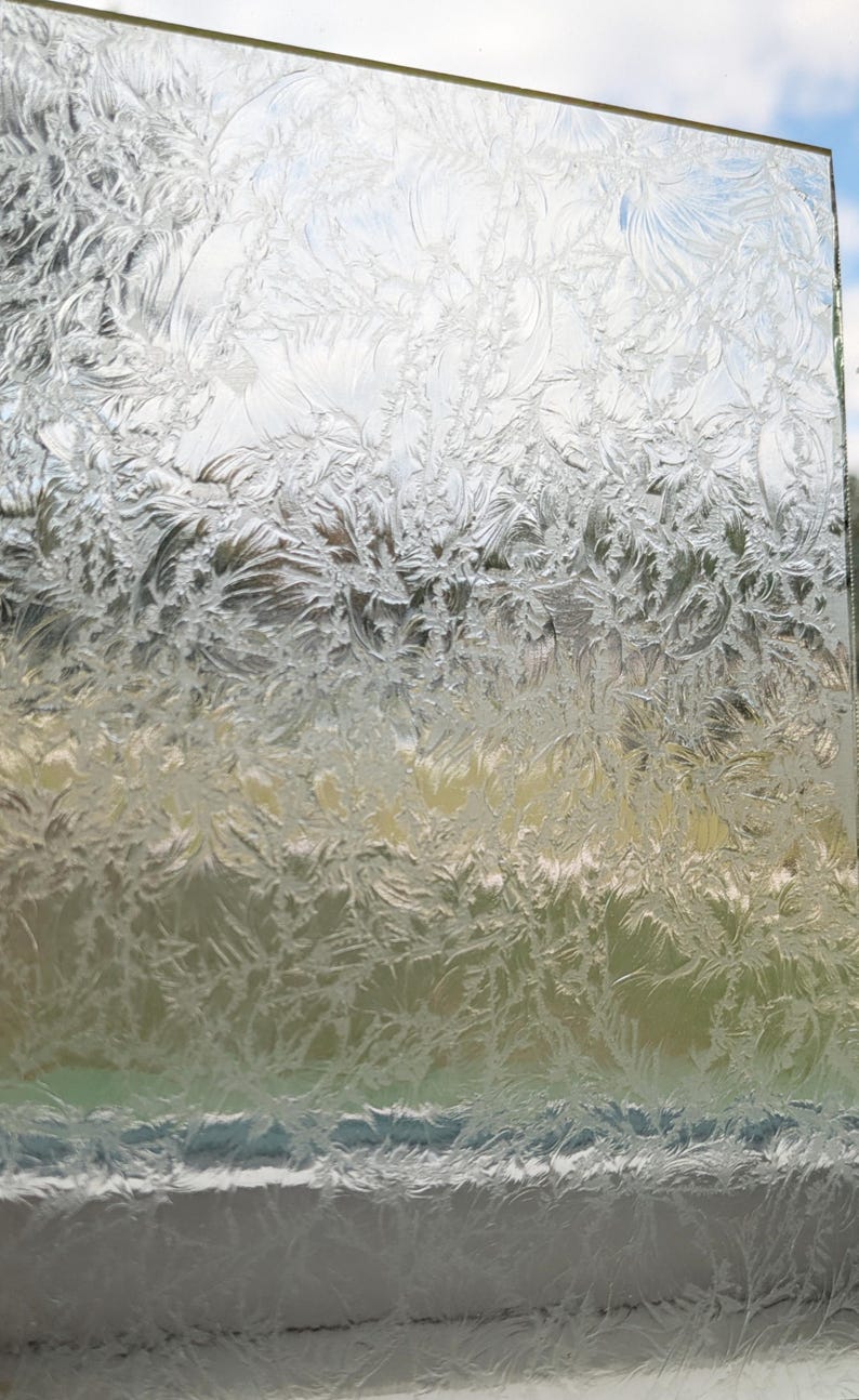 May include: A clear glass panel with a frosted, textured surface. The intricate pattern resembles delicate ice crystals or frost, with a mix of light and shadow. The background shows a glimpse of the sky and other colors.