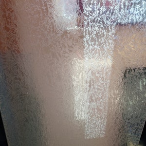 May include: A clear, textured glass panel with a frosted floral pattern. The design features delicate, repeating botanical elements, creating a subtle, elegant aesthetic. The glass has a reflective quality, with light and shadow playing across its surface.