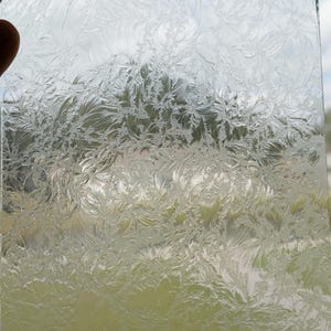 May include: Clear glass panel with a frosted, floral-like pattern. The design resembles delicate ice crystals or frost. The panel is held up against a background of green foliage and a blue sky, creating a contrast between the clear glass and the natural elements.