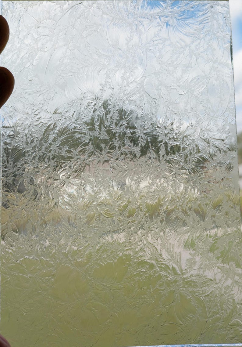 May include: Clear glass panel with a frosted, floral-like pattern. The design resembles delicate ice crystals or frost. The panel is held up against a background of green foliage and a blue sky, creating a contrast between the clear glass and the natural elements.