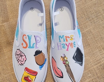 May include: A pair of white canvas slip-on shoes with colorful hand-painted designs. One shoe says "SLP" and has a brain, playdough, and a character from the movie "Toy Story". The other shoe says "Mrs. Hayes" and has a lipstick, a wand, and a graduation cap.