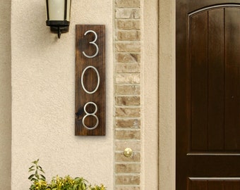 May include: A wooden house number sign with the number 308 in silver metal lettering. The sign is mounted on a brick wall next to a brown wooden door.