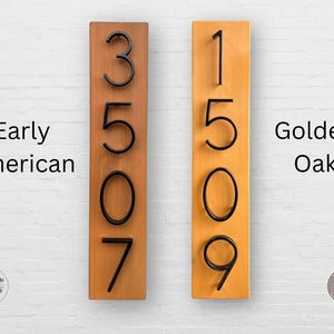 May include: Two wooden house number signs with black numbers. The sign on the left is labeled "Early American" and the sign on the right is labeled "Golden Oak".