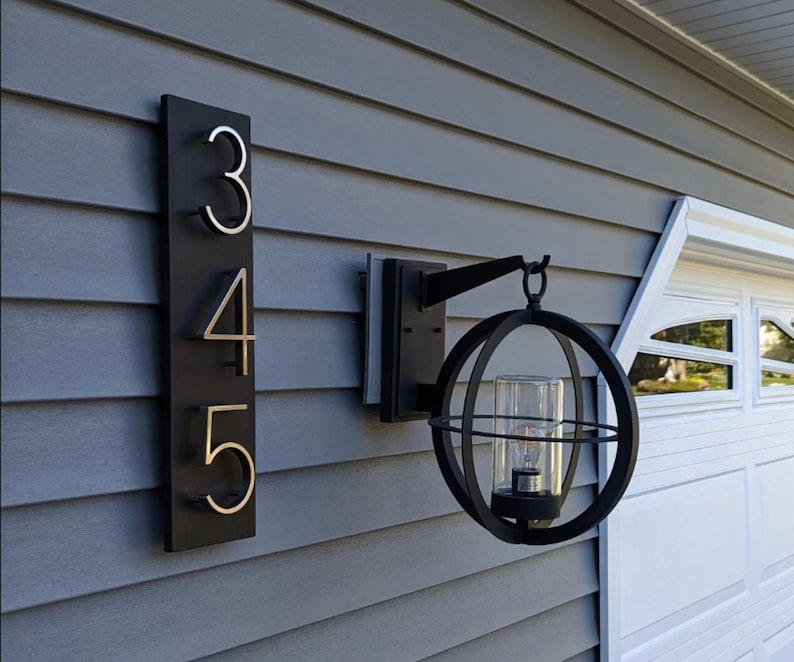 May include: A black metal house number plaque with the numbers 345 in silver on a gray siding background. A black metal globe-shaped outdoor light fixture with a glass globe is mounted on the siding.