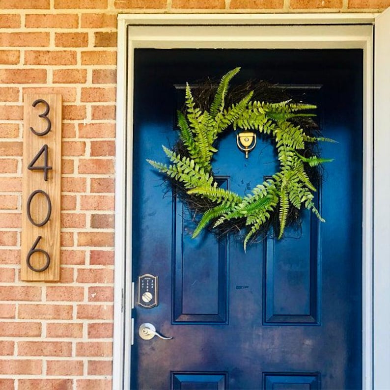 May include: A blue front door with a black metal house number plaque that reads "3406". A green fern wreath hangs on the door. A gold door handle and a digital door lock are visible.