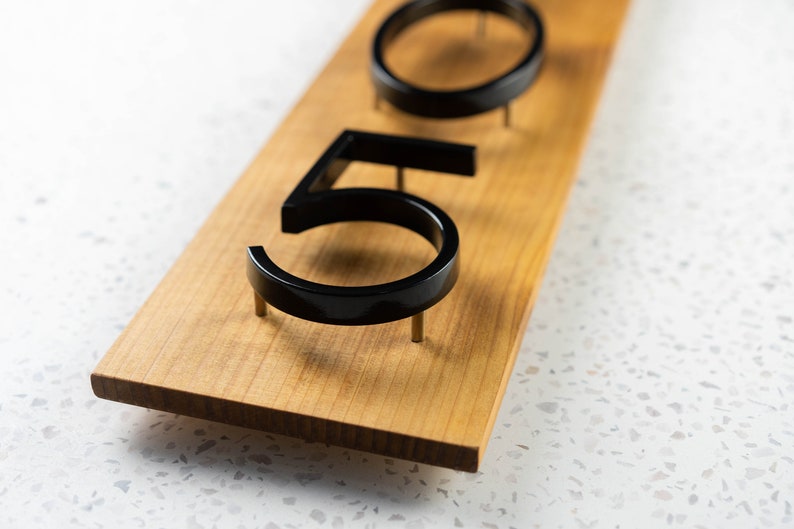 May include: Close-up of a wooden sign with black numbers "50" attached with nails. The sign is on a white speckled surface.