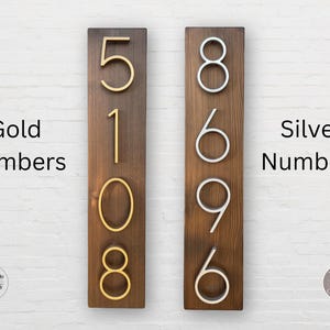 May include: Two wooden signs with house numbers. The sign on the left has gold numbers 5108. The sign on the right has silver numbers 8696. Both signs have a dark brown wood background.