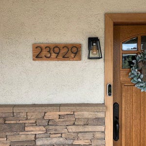 May include: A wooden house number sign with black numbers reading "23929" mounted on a white wall next to a black light fixture and a doorbell.