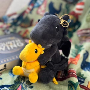 May include: A black plush dragon with a gold keychain clasp hugging a small yellow plush creature. The toys are on a green blanket with a floral and plant pattern. A book is partially visible in the background.