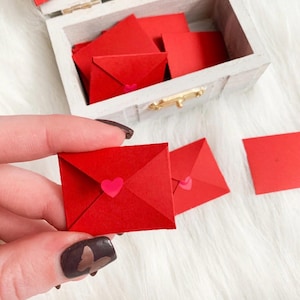 May include: A small, white wooden box with a hinged lid, filled with red paper squares and miniature red envelopes. Each envelope has a pink heart sticker. One envelope is held in a hand.
