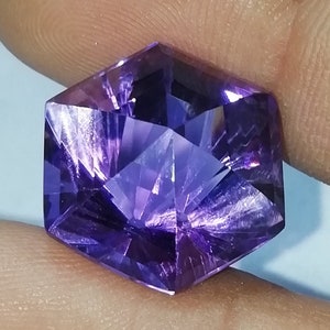 May include: A faceted, hexagonal amethyst gemstone. The gem is a deep purple color with intricate internal cuts that reflect light. The gemstone is held between two fingers, showcasing its shape and color.