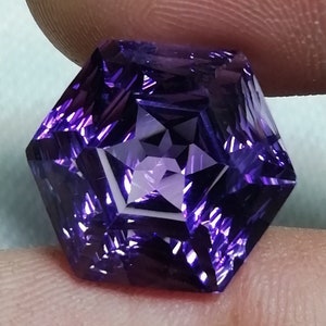 May include: A faceted, hexagonal gemstone with a deep purple hue. The gem's intricate cuts create a star-like pattern in the center, reflecting light. The stone is held between fingers, showcasing its size and detail.