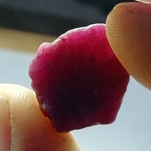 May include: A deep red, faceted gemstone with a rough, irregular shape. The gemstone is held between two fingers.