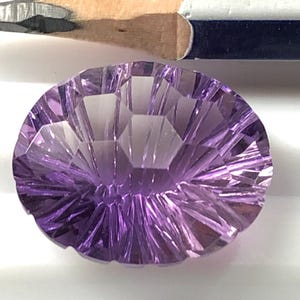 May include: An oval-shaped, faceted purple gemstone with a radial, sunburst-like cut pattern. The gemstone is positioned directly below a sharpened wooden pencil, which serves as a visual reference for the scale of the stone.