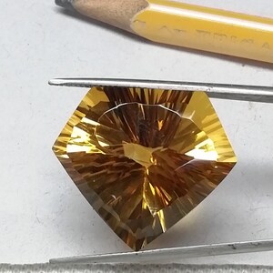 May include: A golden yellow gemstone with a square shape and a starburst pattern. The gemstone is held in place by a pair of tweezers.